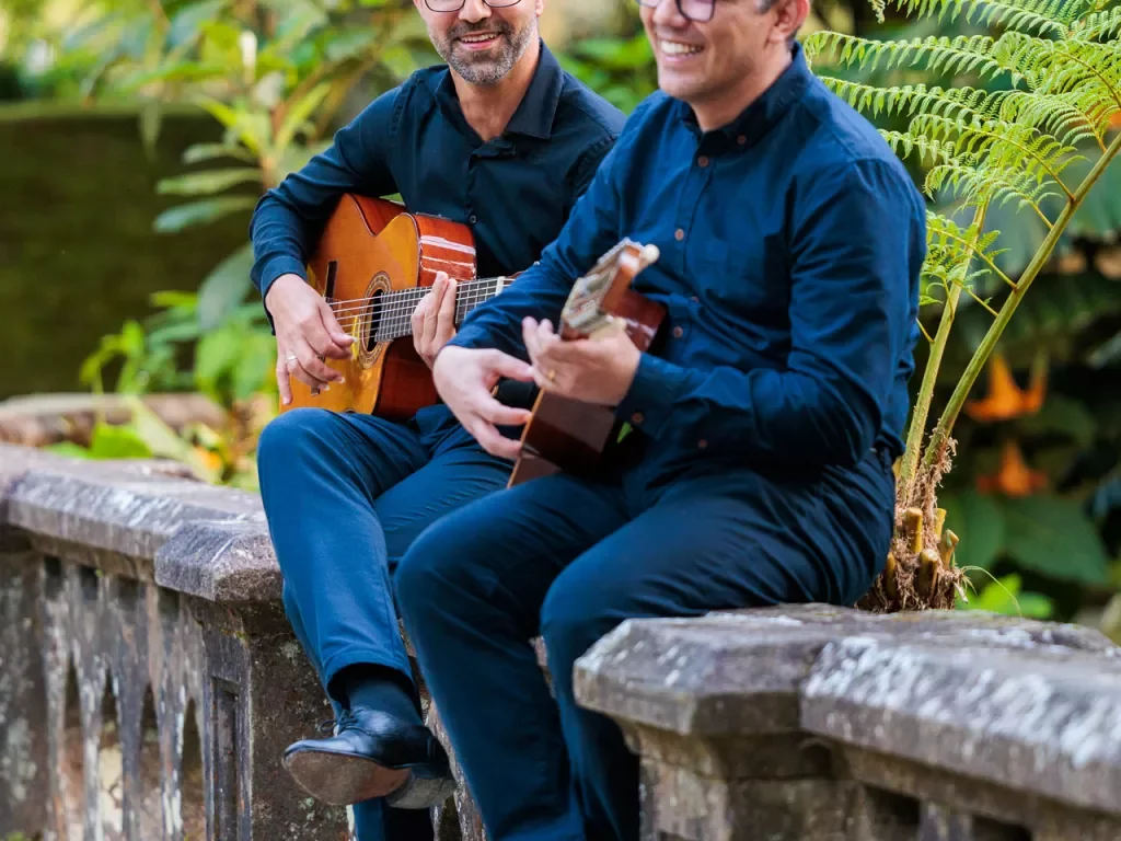 Two men sitting on a stone fence playing guitars and smiling
