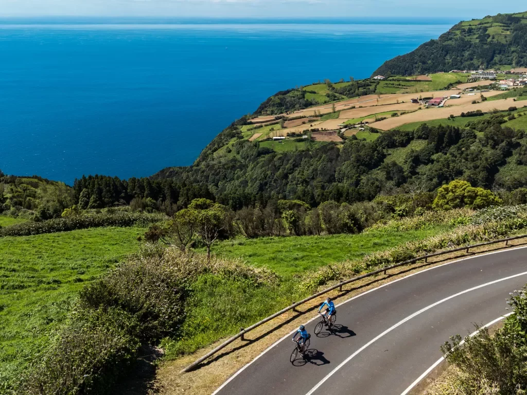 Two people riding their bikes on an asphalt road with a large valley and ocean in the background