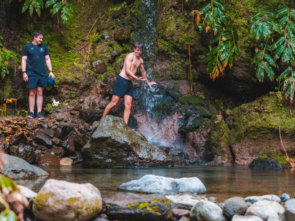 Two men standing on wet rocks under a waterfall, preparing to jump into a lake