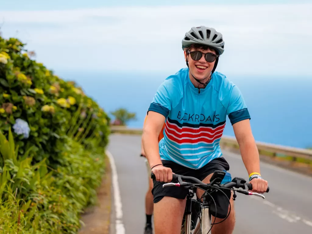 Man with a blue jersey and sunglasses riding a bike on a road next to the ocean