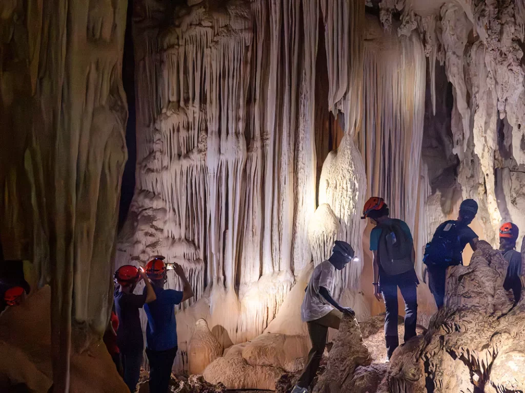 People with hard hats and headlamps exploring a cave with lots of stalactites
