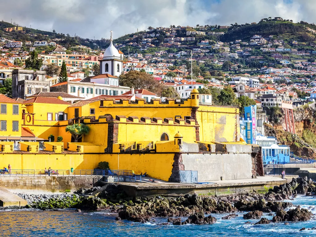 Yellow building by the coast of an ocean, with a hill of houses in the dstance