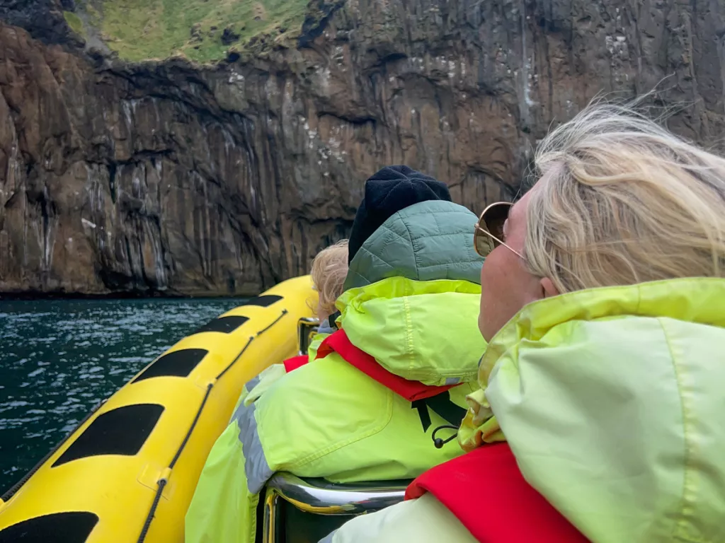 Group of people on a yellow raft, wearing green jackets going towards a cliff