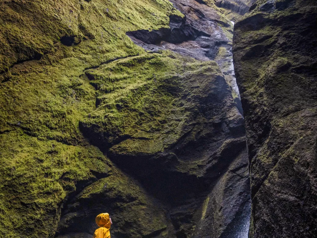 Person in a yellow jacket standing on a rock, looking up to a large cliff