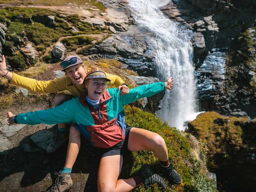 Two women with their arms open on a cliff with a waterfall below