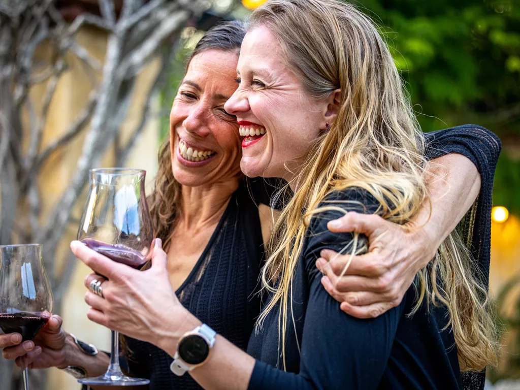 Two women smiling and laughing with arms around their shoulders, holding wine glasses