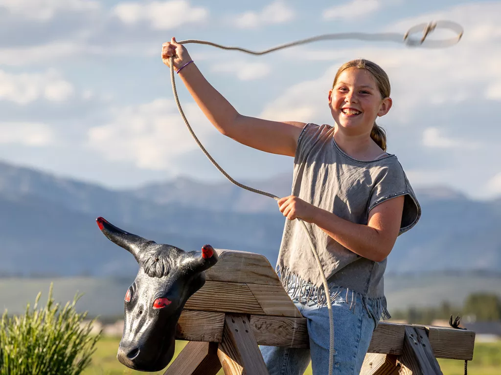 Girl riding a wooden bull prop while swinging a rope