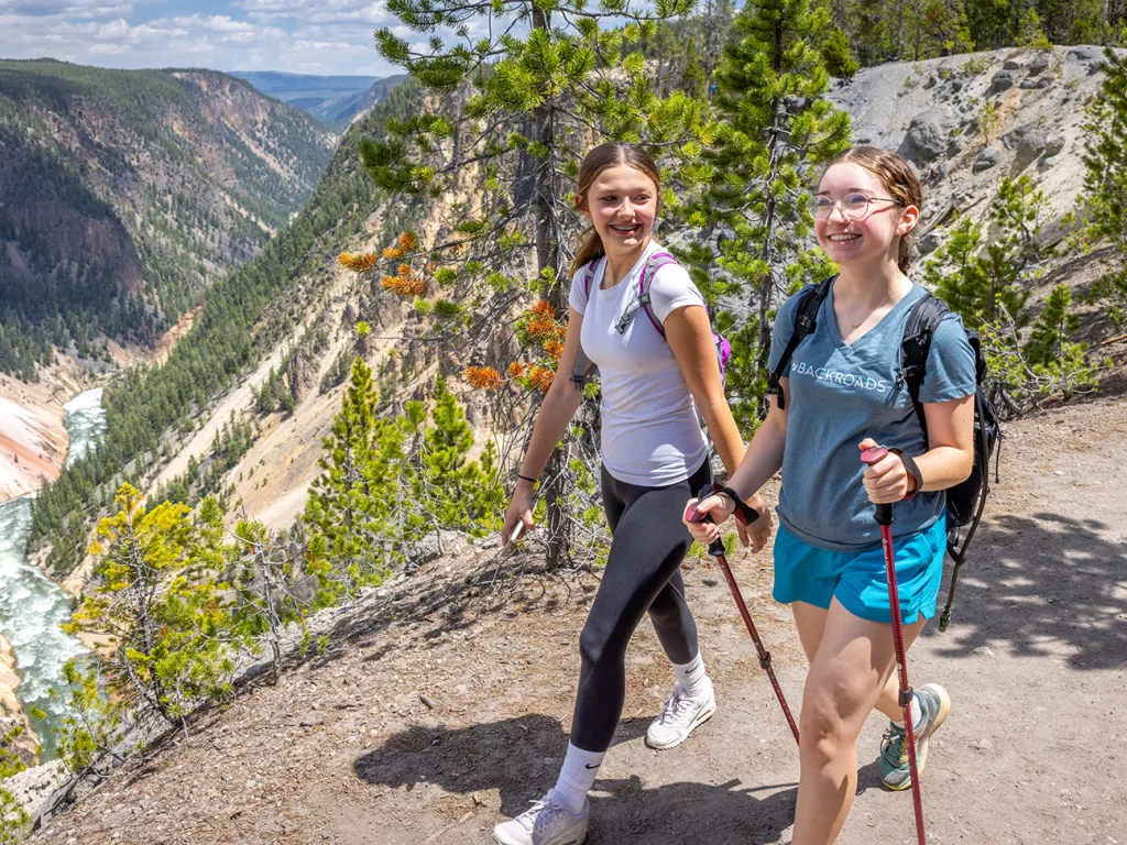 Two girls smiling while hiking, with large mountains and a river in the distance