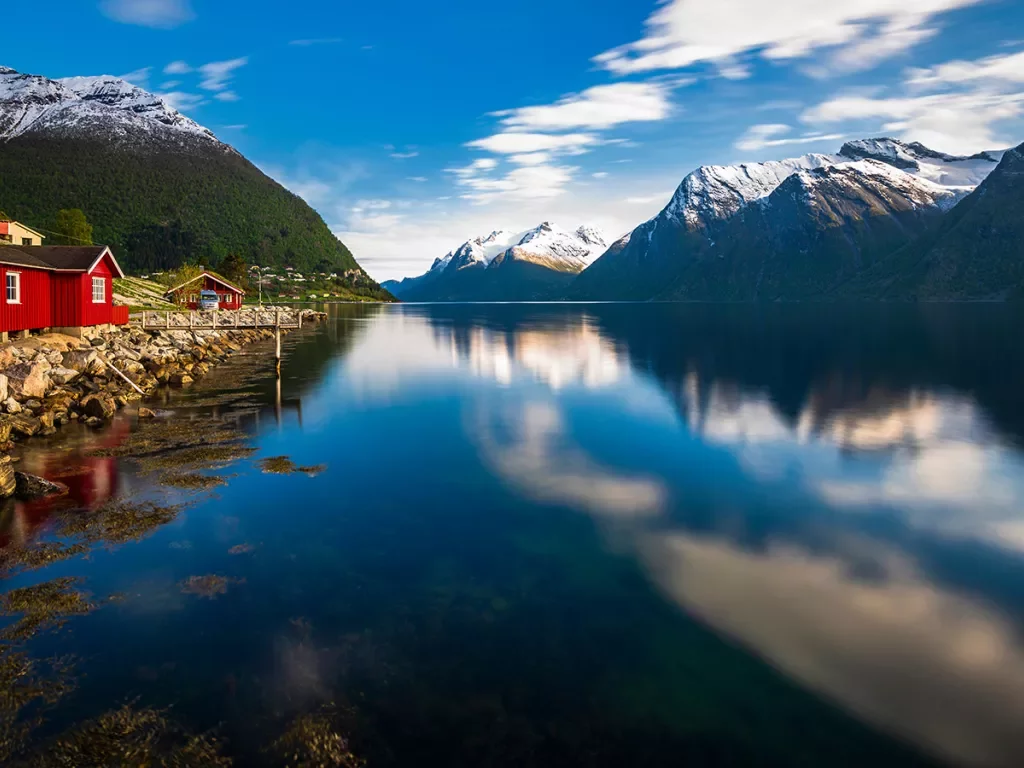 Red house next to an open lake and snowcapped mountains in the background