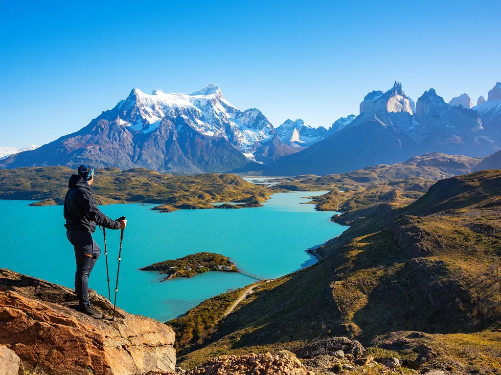 Person with walking poles on top of a rock looking out to a lake and snow-capped mountains