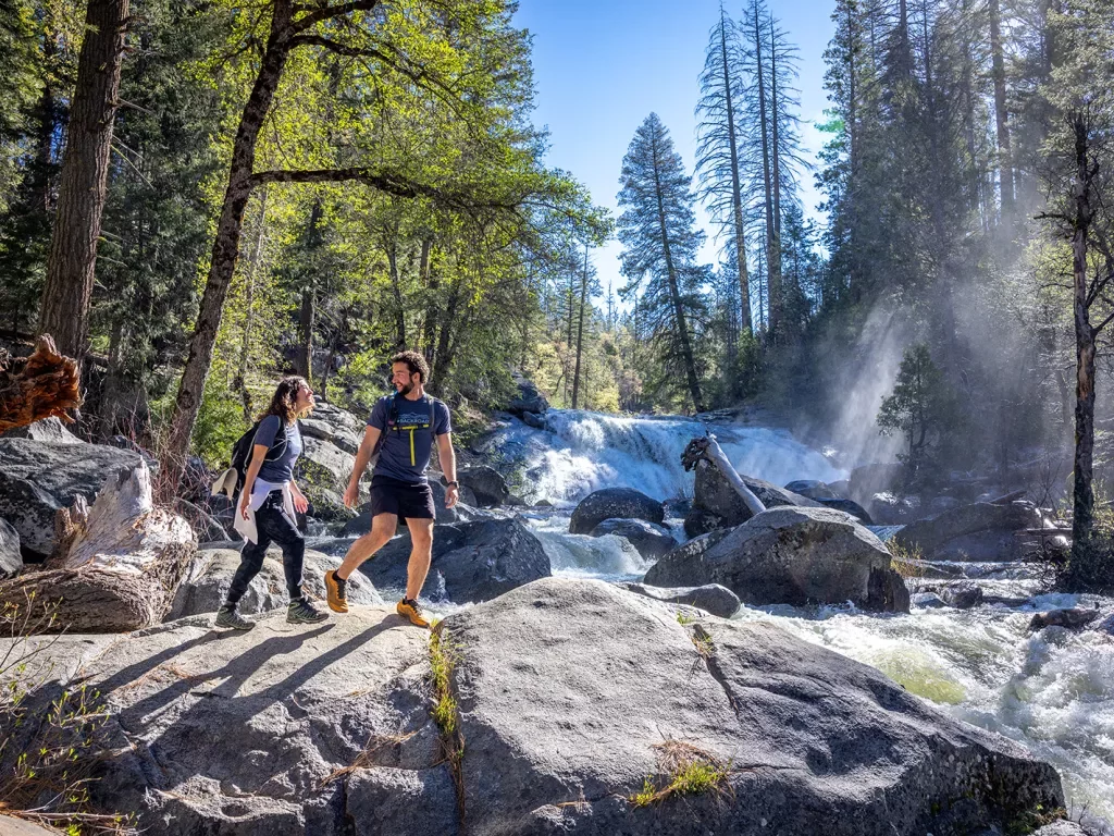 Two hikers cross rocks under a waterfall