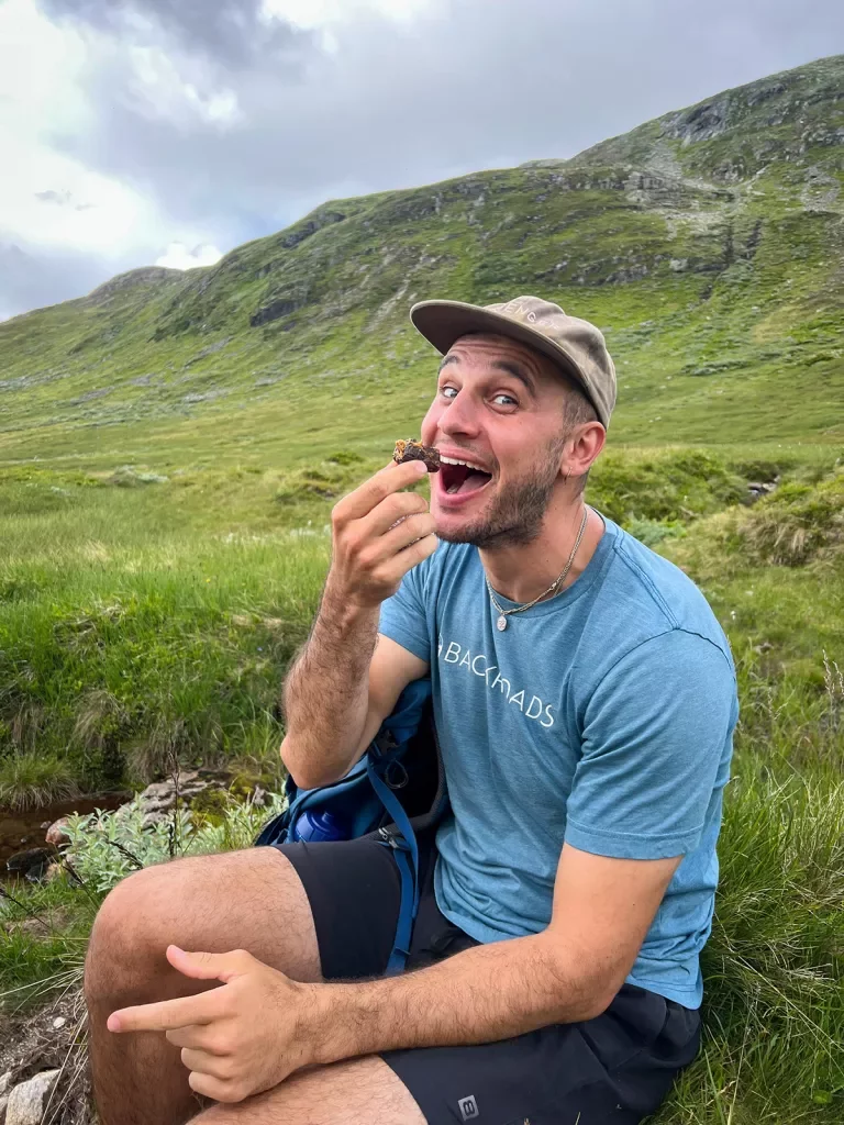 Man sitting in a grassy field with his mouth open eating a snack